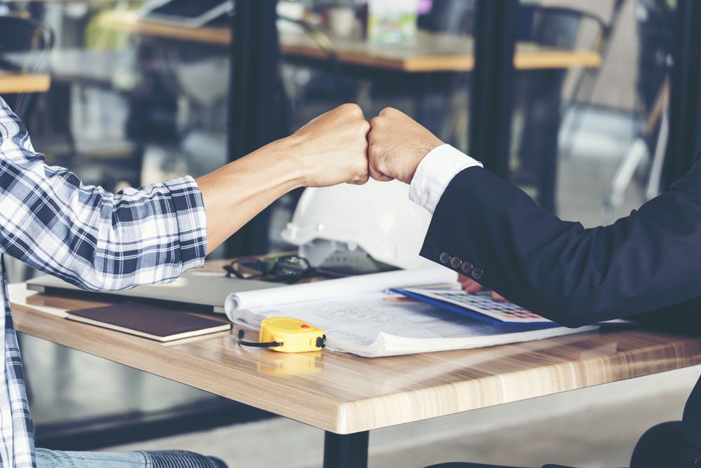 Two men sitting in a meeting space fist bumping each other