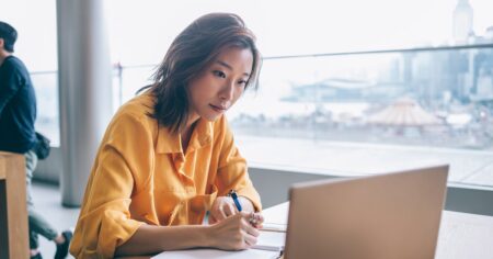 woman in yellow shirt making notes and looking at screen display at desk